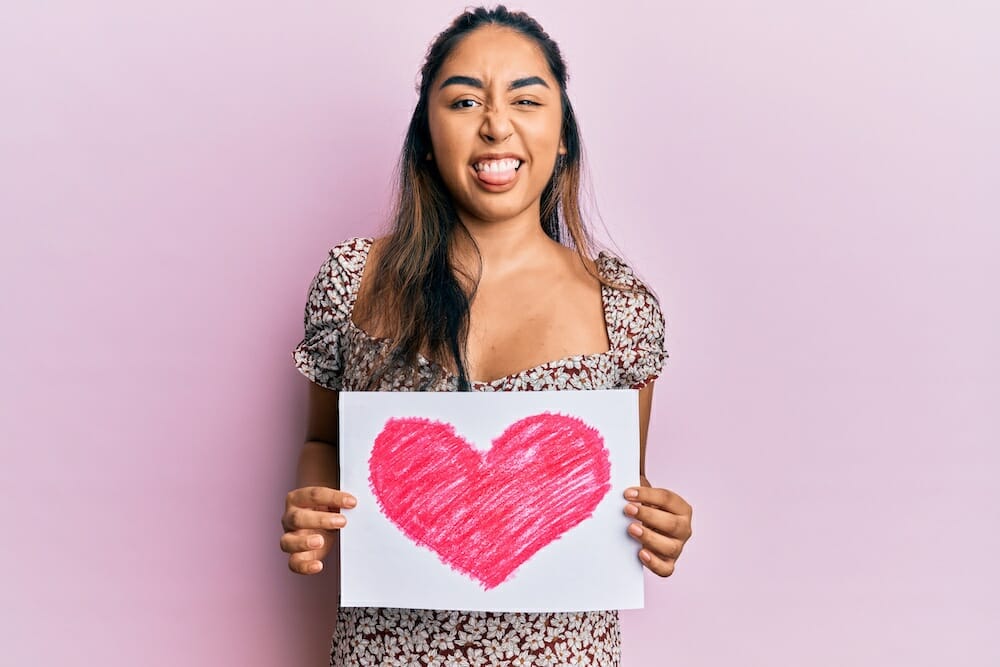 Young latin woman holding heart draw sticking tongue out happy with funny expression.